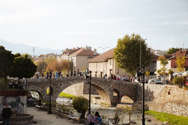 Photo by Sabina Kallari Scenic view of a historic stone bridge over a river in Prizren, Kosovo, with people walking and a charming urban backdrop.